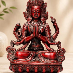 May include: A deep red, four-armed deity statue seated in a cross-legged position on a lotus base. The figure has hands pressed together in a prayer gesture, wearing an ornate crown and jewelry, with flowing garments and a dark, weathered finish.