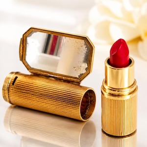 May include: A vintage gold lipstick case with a built-in mirror, next to a gold lipstick tube with a bright red lipstick. The case is cylindrical with vertical ribbed detailing. The mirror is rectangular with an octagonal gold frame.