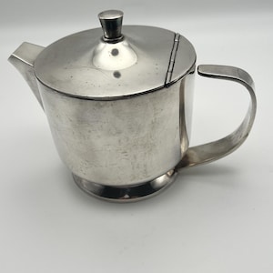 May include: A silver-toned metal teapot with a smooth, polished finish. The teapot features a hinged lid with a small knob handle, a curved spout on the left side, and a C-shaped handle on the right. The base is slightly flared and circular.