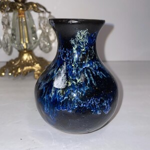 Può includere: Un piccolo vaso in ceramica con smalto blu scuro e nero. Il vaso ha un corpo arrotondato e un bordo leggermente svasato. Lo smalto presenta un effetto marmorizzato con accenti blu chiaro e gialli. Alto circa 10 cm.