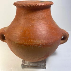 May include: A terracotta-colored earthenware vessel with a wide, rounded body and a narrow neck. The rim features a textured, notched edge, and two small, integrated handles are positioned on opposite sides of the midsection of the pot.