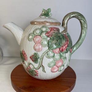 May include: White ceramic teapot with a textured basketweave pattern. The teapot is adorned with a grape cluster design in shades of green, pink, and red. The handle and spout are green, and the lid features a green leaf. The teapot sits on a round wooden stand.