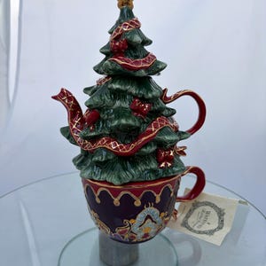 May include: A ceramic teapot shaped like a Christmas tree with a dark green finish, gold star topper, and red ribbon accents. The base is a deep purple teacup with gold trim and intricate patterns. A Waterford Holiday Heirlooms tag is attached.