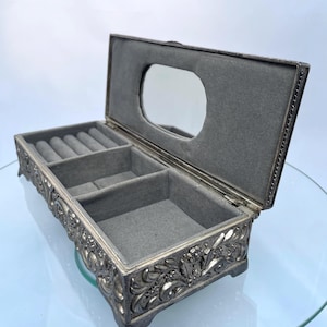 Puede incluir: Un joyero rectangular de tono plateado con un patrón floral en relieve en el exterior. La tapa está abierta, revelando un interior forrado de terciopelo gris con una sección para anillos, dos compartimentos abiertos y un espejo ovalado.