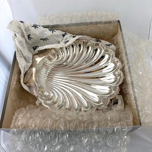 May include: A polished silver-toned serving dish shaped like a large scallop shell with deep, radiating fluted grooves. The dish is nestled inside a square cardboard box lined with clear bubble wrap and white tissue paper featuring a small dark logo.