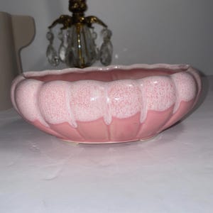 Könnte beinhalten: Ein rosa Keramik-Pflanzgefäß mit gewelltem Rand und gesprenkelter Oberfläche. Der ovale Pflanztopf steht auf einer weißen Oberfläche. Ein Kerzenhalter aus Messing und Kristall befindet sich im Hintergrund.