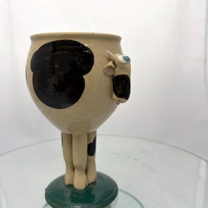 May include: A ceramic goblet featuring a cream-colored body with black spots and a sculpted cow head attached to the side. The base is a solid dark green circle, and the stem is formed by four sculpted legs that connect the bowl to the base.