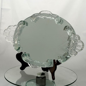 May include: An oval-shaped glass mirror featuring a decorative, clear glass border with intricate, molded fan-like patterns. The mirror is supported by a dark brown, multi-pronged stand, showcasing a reflective surface and detailed glass edges.