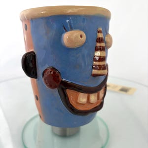 May include: A ceramic mug featuring a sculpted face with a blue surface, a striped nose, and a wide mouth showing teeth. The side of the mug is pink with black polka dots, and it has a dark brown ear-like protrusion on the left side of the face.