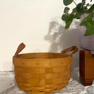 May include: A round, woven wooden splint basket with a light brown finish. The basket features a reinforced rim secured with small metal tacks and two tan leather handles attached to opposite sides. The weave pattern is horizontal and vertical.