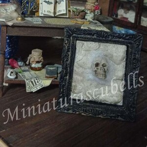 May include: A miniature witch doll in a blue dress and hat stands in a detailed room setting. A table displays a book with bat illustrations, jars, and a framed skull art piece. The scene is a whimsical, handcrafted diorama.