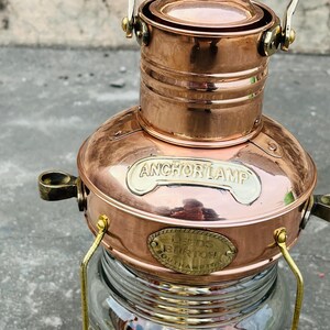 Op de afbeelding: Koperen ankerlamp met een heldere glazen basis. De lamp heeft de tekst "ANCHOR LAMP" en een plaatje met "LEEDS BURTON SOUTHAMPTON". Het heeft een handvat en messing accenten. Een nautisch item.