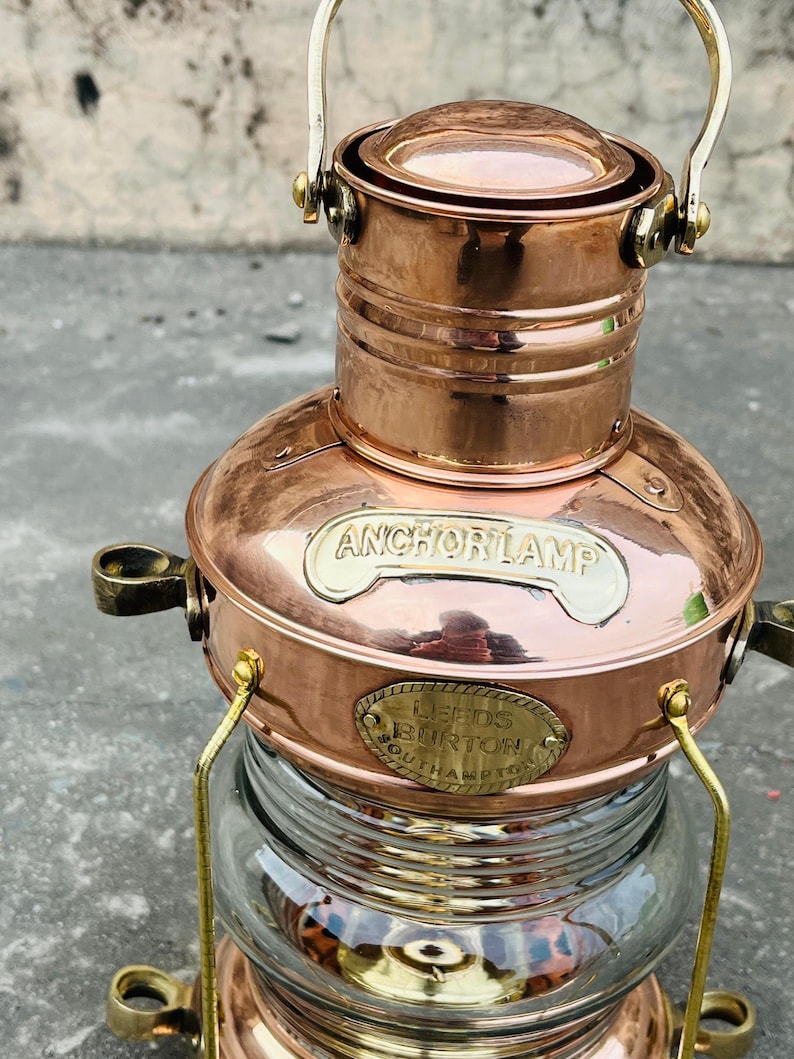 Op de afbeelding: Koperen ankerlamp met een heldere glazen basis. De lamp heeft de tekst "ANCHOR LAMP" en een plaatje met "LEEDS BURTON SOUTHAMPTON". Het heeft een handvat en messing accenten. Een nautisch item.