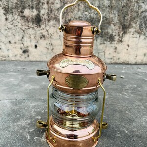 Op de afbeelding: Een nautische lantaarn van koper en messing met een heldere glazen bol. De lantaarn heeft een handvat en decoratieve messing accenten. De tekst op de lantaarn luidt "Nautical" en "Leeds, England".