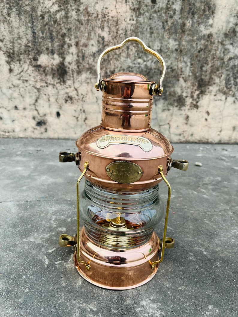 Op de afbeelding: Een nautische lantaarn van koper en messing met een heldere glazen bol. De lantaarn heeft een handvat en decoratieve messing accenten. De tekst op de lantaarn luidt "Nautical" en "Leeds, England".
