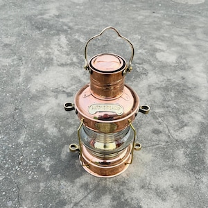 Op de afbeelding: Een koperkleurige ankerlamp met een heldere glazen lens. De lamp heeft een handvat en is gemarkeerd met "ANCHOR LAMP" en "LIBERTY LONDON". Het ontwerp is klassiek en nautisch.
