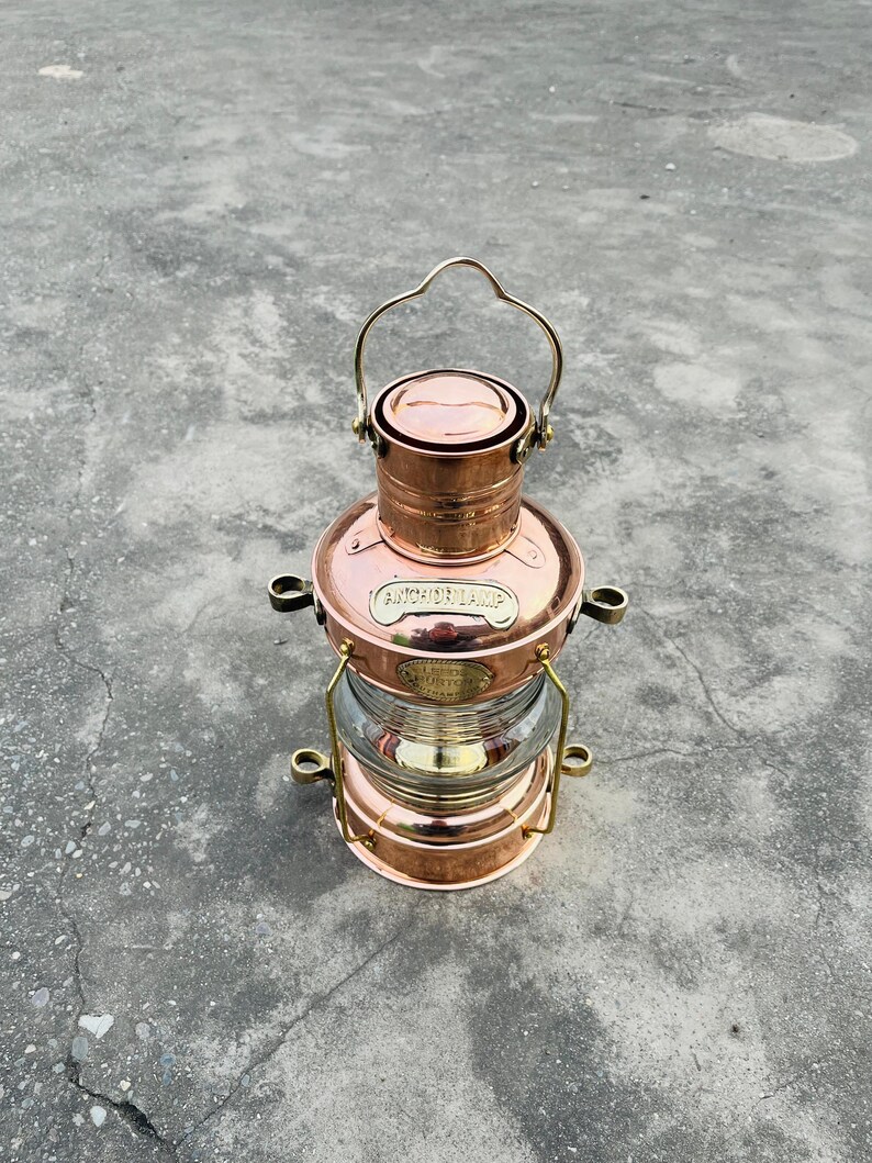Op de afbeelding: Een koperkleurige ankerlamp met een heldere glazen lens. De lamp heeft een handvat en is gemarkeerd met "ANCHOR LAMP" en "LIBERTY LONDON". Het ontwerp is klassiek en nautisch.