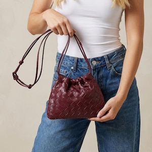 May include: A burgundy woven leather crossbody bag with a drawstring closure. The bag has a long, thin strap and is held by a person wearing a white top and blue jeans.