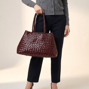 May include: A burgundy leather tote bag with a woven pattern. The bag features braided handles and a rectangular shape. The bag is held by a person wearing a grey cardigan and navy trousers.