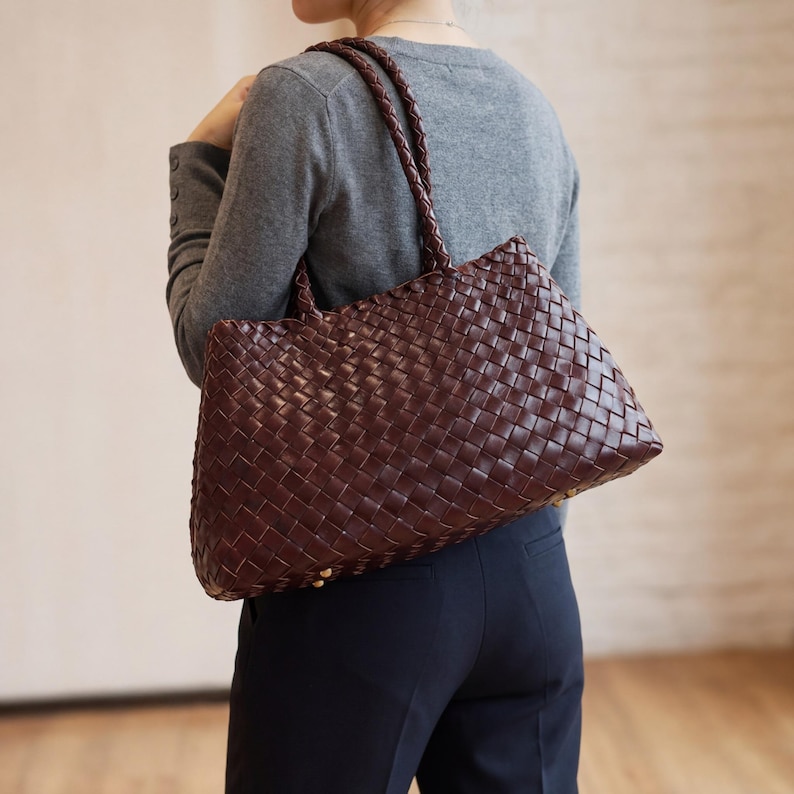 May include: A dark brown woven leather tote bag with braided handles. The bag is rectangular with a woven pattern and gold-coloured rivets at the base. The bag is carried over the shoulder.
