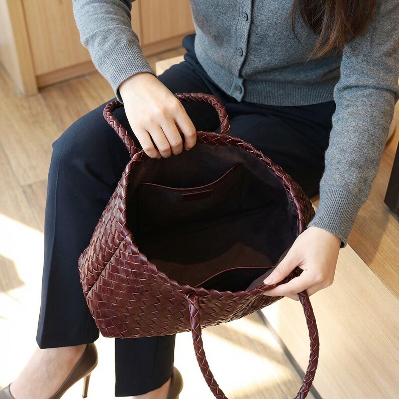 May include: A burgundy woven leather tote bag with braided handles. The bag has an open top, revealing a dark interior with pockets. The bag is held by a person sitting down.