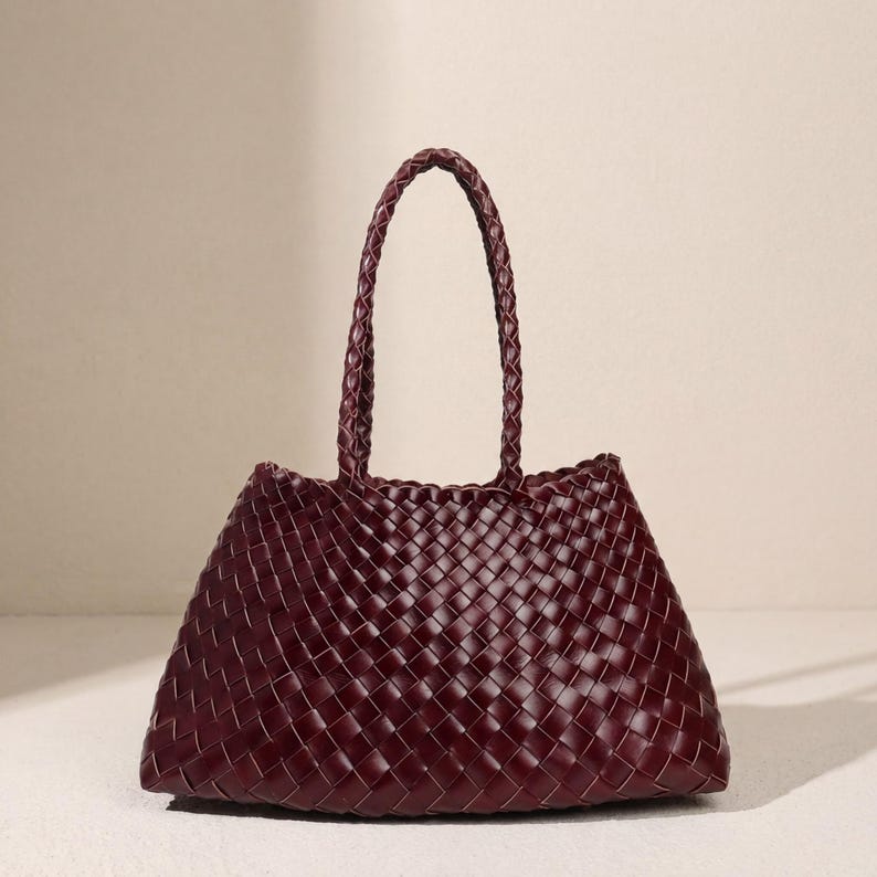 May include: A burgundy tote bag with a woven design. The bag has a trapezoid shape and a braided handle. The woven pattern is visible on the body of the bag and the handle. The bag is made of a soft material.