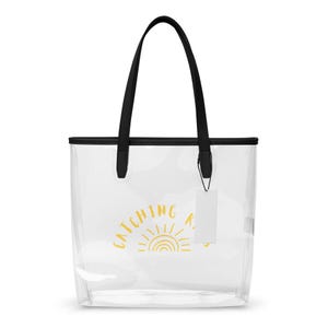 May include: A clear tote bag with black shoulder straps and black trim. The front features a yellow graphic of a sun with the text "CATCHING RAYS" arched above it. A small white rectangular tag hangs from one of the black strap attachments.