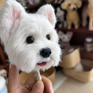 Op de afbeelding: Een witte West Highland Terrier hondenhoofdsculptuur met zwarte ogen en neus, en roze binnenoren. De tong van de hond is zichtbaar. De sculptuur wordt door een hand vastgehouden, met andere hondenbeeldjes op de achtergrond.