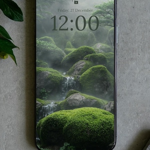 Pode incluir: Um smartphone com moldura preta exibindo um papel de parede digital de uma paisagem rochosa enevoada coberta de musgo com uma pequena cachoeira. A tela mostra a hora 12:00 e a data sexta-feira, 27 de dezembro, com um ícone de cadeado.