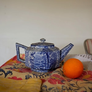 以下が含まれることがあります： 風車、木、人物が描かれた風景デザインの青と白のセラミックティーポット。 ティーポットは、明るいオレンジ色の果物の隣にある花柄の布の上に置かれています。 ティーポットは幾何学的なハンドルと注ぎ口を備えています。