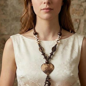 May include: A brown beaded necklace with a large heart-shaped pendant. The necklace features wooden beads, shell accents, and a tassel. The pendant has a natural wood grain pattern. The woman is wearing a cream-colored dress.