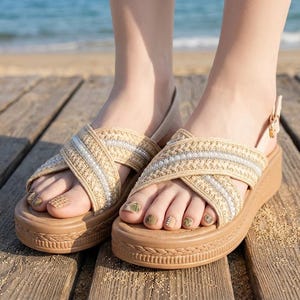 May include: Tan platform sandals with woven straps and a cross-over design. The straps are embellished with silver and white beads. The sandals have a buckle closure and are worn on a wooden deck.