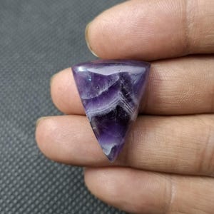 May include: A triangular amethyst gemstone with a polished surface. The stone displays a gradient of purple hues, with white and dark purple bands. The gemstone is held in a hand against a dark gray background.