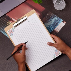 May include: A wooden clipboard with a silver clip holding lined paper. The paper has floral corner designs in pink and blue. A black pen is held over the paper. The word "DATE" is visible at the top of the page.