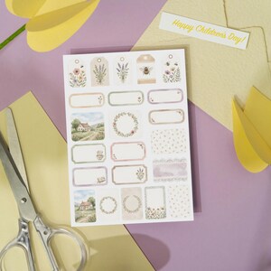 May include: A sheet of decorative stickers with floral and landscape designs, including tags and labels. A pair of scissors and a paper tulip are on a purple surface. A yellow envelope and a card that says "Happy Children's Day!" are also present.