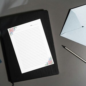 May include: A stationery set featuring a black notebook, lined paper with floral corner accents, a light blue envelope, and a pencil. The paper has a space for the date at the top. The floral accents include pink and blue flowers.