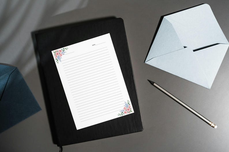 May include: A stationery set featuring a black notebook, lined paper with floral corner accents, a light blue envelope, and a pencil. The paper has a space for the date at the top. The floral accents include pink and blue flowers.