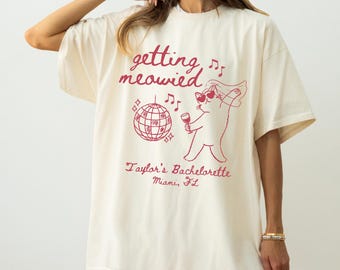 Op de afbeelding: Een off-white t-shirt met korte mouwen en een rode grafische print. Het ontwerp bevat de tekst "getting meowied", een discobal, muzieknoten en een kat met een hartvormige zonnebril en een sluier, die een glas vasthoudt. Tekst: "Taylor's Bachelorette Miami, FL".