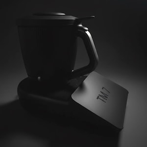 Pode incluir: Um eletrodom&eacute;stico de cozinha preto com uma al&ccedil;a e uma tampa. O aparelho est&aacute; sobre uma base preta e tem uma etiqueta retangular com o texto "TM 7". A imagem &eacute; em preto e branco.