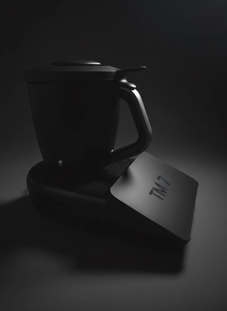 Pode incluir: Um eletrodom&eacute;stico de cozinha preto com uma al&ccedil;a e uma tampa. O aparelho est&aacute; sobre uma base preta e tem uma etiqueta retangular com o texto "TM 7". A imagem &eacute; em preto e branco.