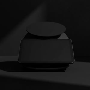 Pode incluir: Uma balan&ccedil;a de cozinha digital preta com uma base retangular e um topo redondo. A balan&ccedil;a est&aacute; sobre um fundo escuro com um efeito de holofote, destacando seu design elegante. &Eacute; usada para medir ingredientes.