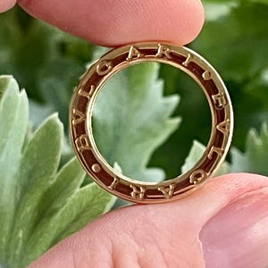 May include: A gold ring with the word "BVLGARI" engraved around the outer edge. The ring is held up against a blurred green background. The ring is circular with a hollow centre.