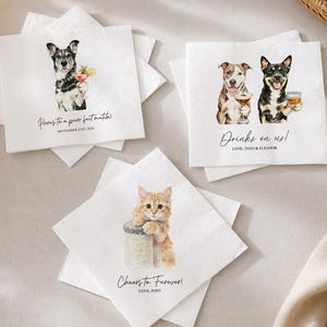 Può includere: Tre tovaglioli di carta bianchi con illustrazioni di animali domestici. Uno mostra un cane con un cocktail e il testo "Here's to a purr-fect match! NOVEMBER 21ST, 2026". Un altro mostra due cani con bevande e "Drinks on us! LOVE, THEO & ELEANOR". Il terzo mostra un gatto e "Cheers to Furever! LOVE, ZOEY".