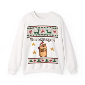 May include: A white long-sleeve sweatshirt featuring a graphic print of a brown beaver wearing a Santa hat, surrounded by yellow stars. The text Yule Log Expert is printed above the beaver, framed by horizontal bands of green and red patterns.