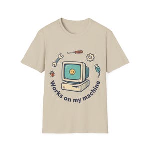 Op de afbeelding: Beige t-shirt met een retro computerontwerp, een lachend gezicht op het scherm en de tekst "Works on my machine". Het ontwerp bevat een moersleutel, schroevendraaier en USB-kabel.