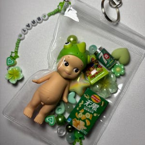 May include: A clear plastic pouch filled with a baby doll wearing a green frog hat, miniature snacks, and green beads. A beaded keychain with the word "BRAT" and flower charms is attached.