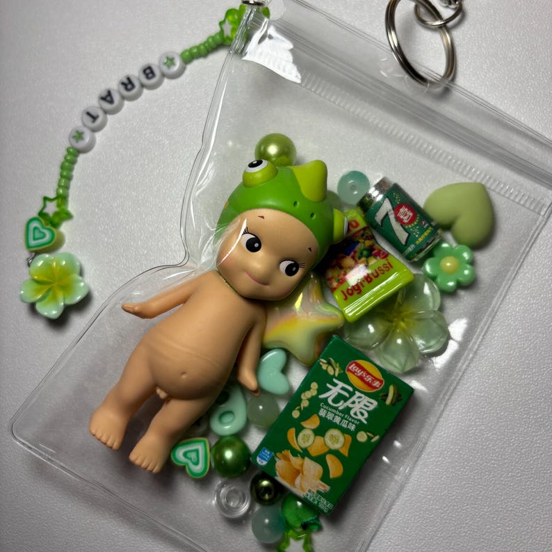 May include: A clear plastic pouch filled with a baby doll wearing a green frog hat, miniature snacks, and green beads. A beaded keychain with the word "BRAT" and flower charms is attached.