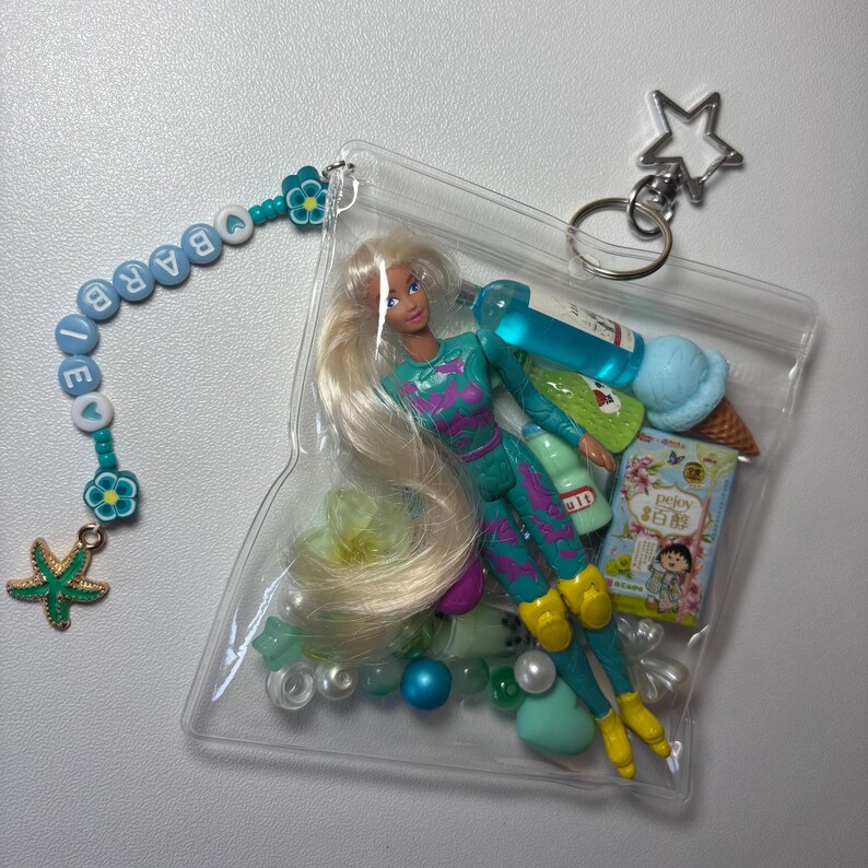 K&ouml;nnte beinhalten: Ein durchsichtiger Kunststoffbeutel mit einer Miniaturpuppe mit langem blondem Haar, einem t&uuml;rkisfarbenen gemusterten Outfit und gelben Stiefeln. Im Inneren befinden sich kleine Spielzeugaccessoires, darunter eine blaue Eiswaffel, eine Trinkflasche und eine Schachtel mit der Aufschrift Pejoy. Eine Perlenkette mit dem Wort BARBIE und einem gr&uuml;nen Seestern-Anh&auml;nger ist am Beutel befestigt, der einen sternf&ouml;rmigen Metallschl&uuml;sselring besitzt.