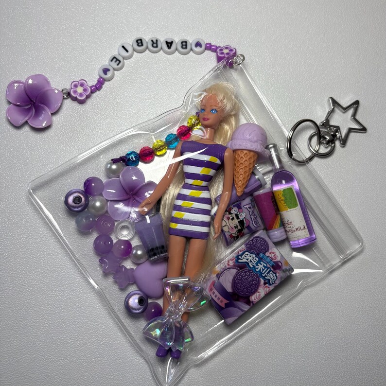 K&ouml;nnte beinhalten: Ein durchsichtiger Kunststoffbeutel mit einer Puppe in einem violett-wei&szlig; gestreiften Kleid, umgeben von Miniaturgegenst&auml;nden wie einer violetten Eist&uuml;te, einer Flasche, einer Snackpackung mit asiatischem Text, Perlen und einem Blumenanh&auml;nger.