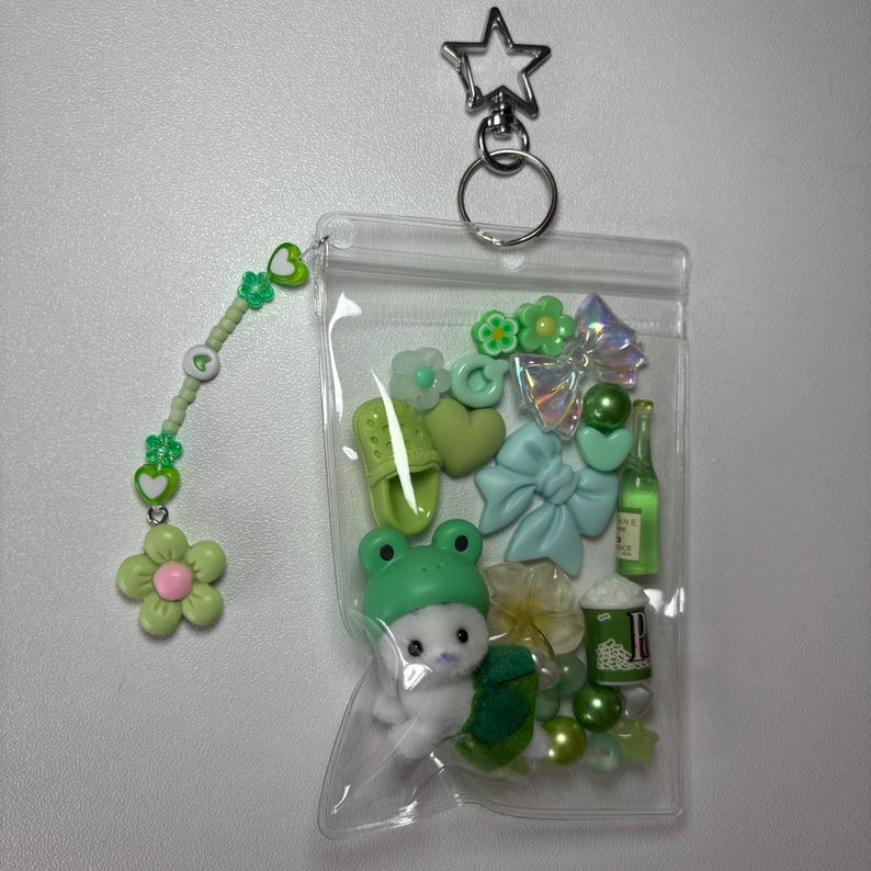 May include: A clear plastic keychain filled with green and white charms, including a frog hat, a bunny, a bow, and a flower. A beaded charm with a flower and heart accents hangs from the side. The keychain has a silver star-shaped clasp.